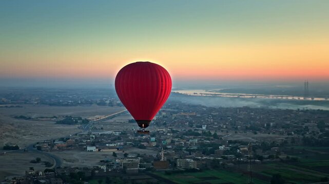 Aerial view of a red hot air balloon floating above the West Bank city layout in Luxor, back-lit by the vibrant orange and blue gradients of a hazy sunrise over the Nile River.