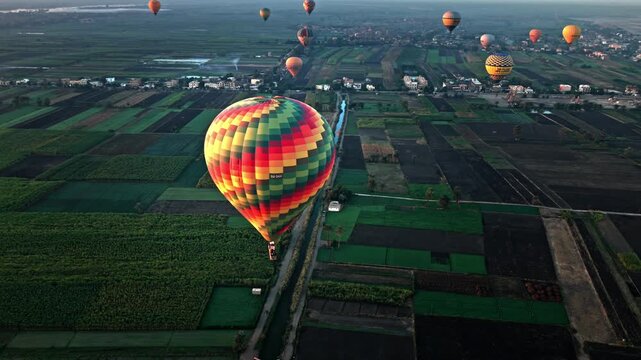 Aerial view focused on a multi-colored patterned hot air balloon gliding low over a patchwork of lush green agricultural fields and irrigation canals on Luxor's West Bank, with other balloons.
