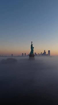 Slow vertical pan drone shot of silhouetted Statue of Liberty, New York. Pre-dawn light, low sea fog, clear sky. Manhattan skyline in background