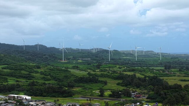 Row of white wind turbines crowns lush green rolling hills above Kahuku coastal community with colorful homes and swaying palm trees under cloudy skies in Hawaii, USA. Aerial drone wide shot.
