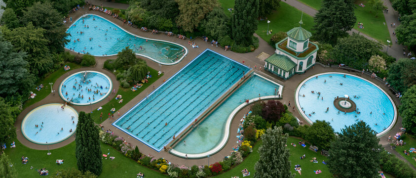 An aerial view of an outdoor public swimming pool complex featuring multiple pools of various shapes and sizes, surrounded by lush green trees and grass.