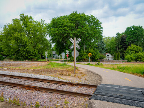 A gravel trail and bike path crosses a single railroad track at a suburban crossing with crossbuck signs under a cloudy sky
