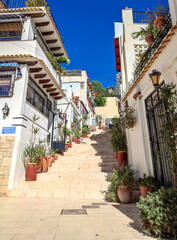 Fototapeta premium Beautiful traditional narrow street staircase in old town Barrio de Santa Cruz Alicante Spain with white houses and terracotta flower pots under clear blue sky