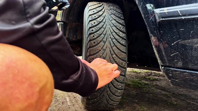 Mechanic crouching near black vehicle and using hand tool to clear debris from tire tread slots in timelapse scene