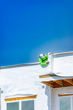 A vertical view of a construction worker wearing a white hard hat, a bright neon green safety shirt, and a safety harness, leaning over the roof edge to install white weather-resistant housewrap barri