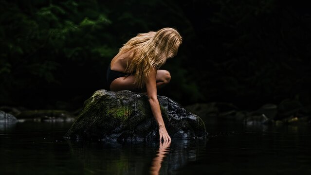 Serene Woman with Long Blonde Hair Sitting on Mossy Rock in Dark Forest River