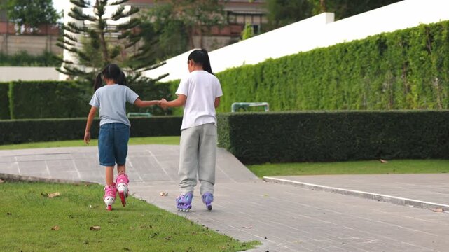 Cute young Asian children girls rollerskating and balancing together inside summer public park green field. Happy classmates enjoying joyful outdoor exercise track process.