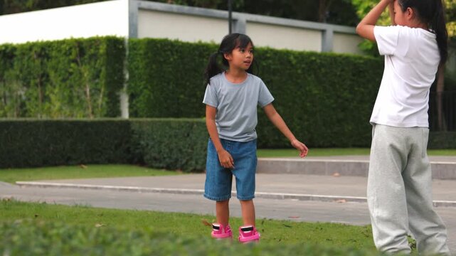 Two happy Asian kids girls learning how to rollerskate together inside green public park courtyard. Cute young classmates sharing friendship outdoor lifestyle morning project.