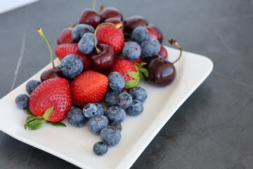 Fresh summer berries mix on white plate side view. Fresh summer berries including strawberries, blueberries and sweet cherries served on a white square plate, top view. © Nadiia