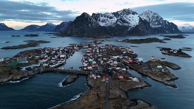 Aerial Drone Panorama of Henningsvaer Fishing Village and Football Stadium, Lofoten, Norway, at blue hour with snowy mountains in the background, Horizontal backwards revealing shot
