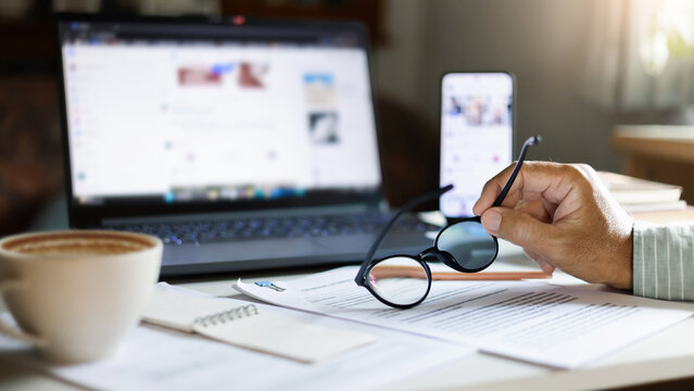 close up shot, man hand holding eyeglasses working online at home with business documents and laptop start new business, E-commerce