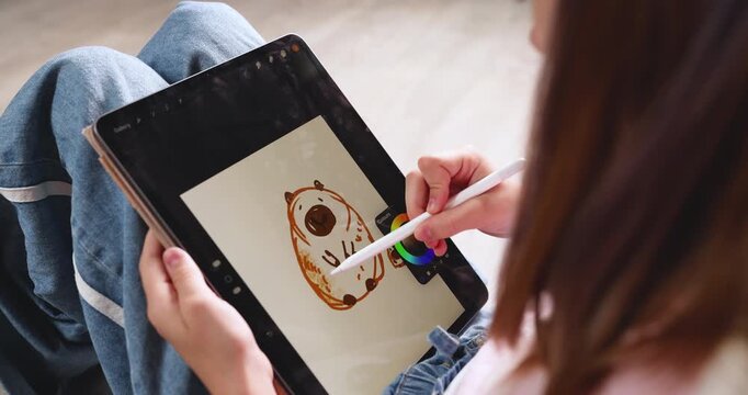 Child wearing pink cap drawing cute cartoon capybara on digital tablet screen with stylus pen. Creative educational graphic design software activity for early child development at home. Real indoor