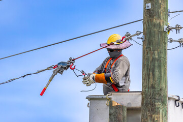 A close-up horizontal view of a utility lineman working from a bucket truck, wearing a hard hat with a sun brim, safety harness, and heavy protective gloves while pulling and splicing an overhead line © Andrei Liashchou