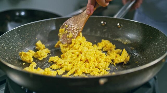 Chef scrambling eggs in frying pan on stove, close up, breakfast preparation, healthy food cooking, culinary arts, kitchen motion footage.