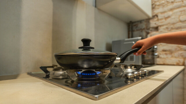 Child's hand holding a frying pan on a gas stove with a blue flame