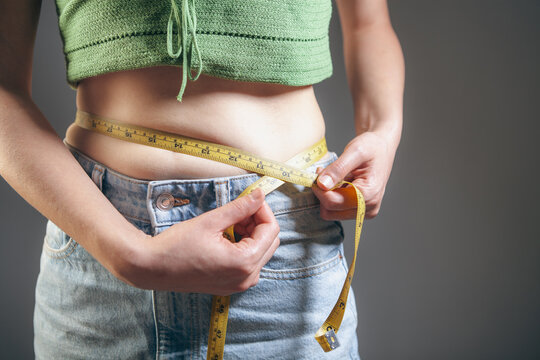 Woman measuring her waist with a tape measure on a gray background.