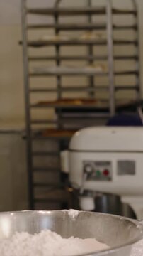 Vertical video: Entering left baker in apron dusting flour at bakery reaching mixing bowl for dough