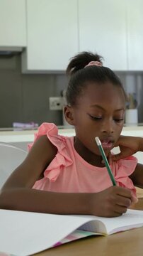 Vertical video: Holding green pencil, girl in pink top writing homework in notebook, mom with mug