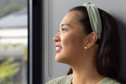 Mid Adult Asian woman gazing out window at home by blind chain, wearing headband, copy space
