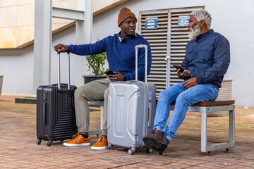 Sitting two men wearing beanie and zip jacket talking at plaza, holding smartphones and suitcases © wavebreak3