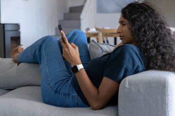 Obraz premium African American woman reclining on light grey sofa, holding smartphone and checking smartwatch