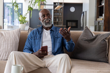 Obraz premium African American man sitting on beige sofa in living room, holding red smartphone and gesturing