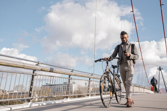Stylish man with a bicycle using smartphone while walking on a bridge