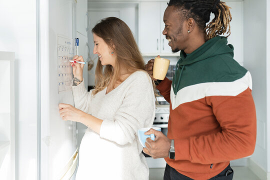 Pregnant woman with man in kitchen crossing date on calendar at home