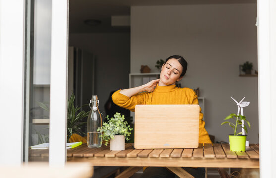 Young freelancer with neckache sitting at desk seen through window