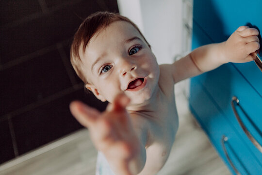 Laughing baby boy holding on to chest of drawers, reaching up
