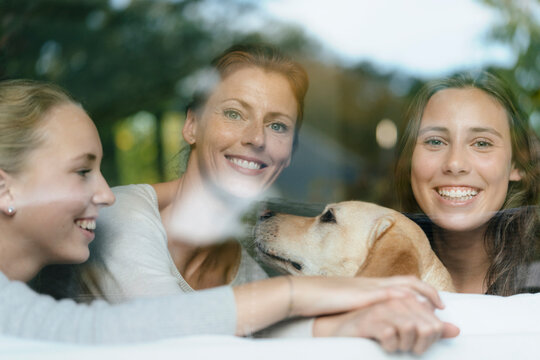 Portrait of happy mother with two teenage girls and dog behind windowpane