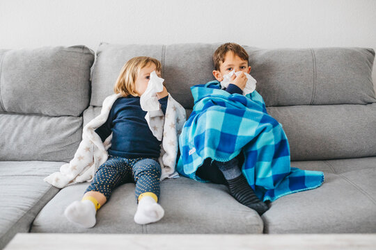 Little girl and her brother sitting side by side on the couch at home blowing noses
