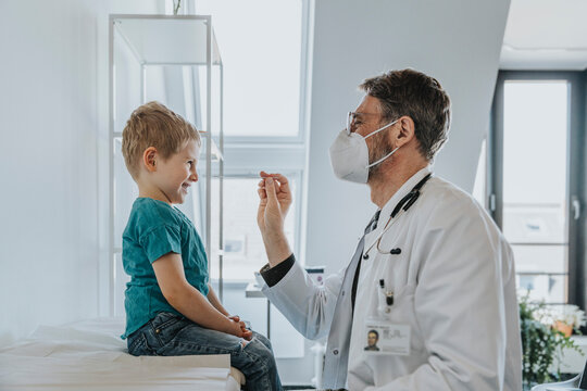 Pediatrician wearing protective face mask taking nasal swab test of boy while standing at clinic