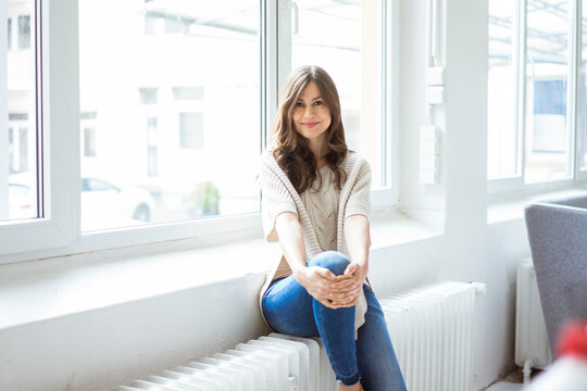 Portrait of smiling woman sitting on heater in bright room with window