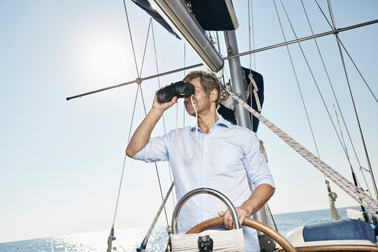 Mature man using binoculars at helm of his sailing boat