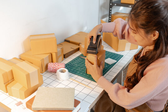 A young woman is carefully assembling cardboard boxes at a neatly organized workspace. The desk is filled with packing materials, tape, and string, reflecting focus and small business activity.