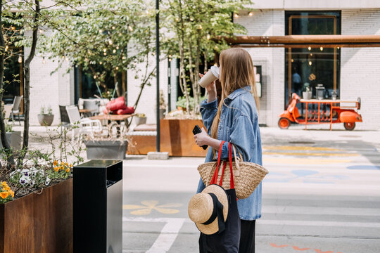 Woman standing outdoors sips beverage while holding smartphone and carrying woven tote bag. Sustainable living, urban lifestyle, eco friendly choices.