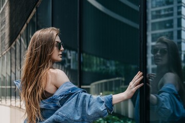Fototapeta premium Woman wearing sunglasses looks at her reflection in glass facade of modern office building. self reflection concept, introspective business professional, identity in modern city.