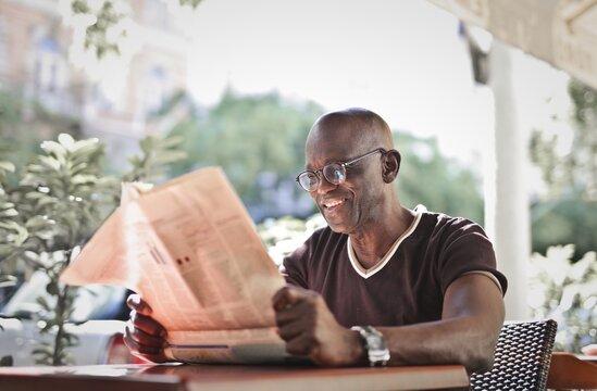 Smiling man with glasses reading newspaper outside