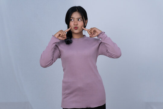 Cute Asian woman in purple shirt poking cheeks and puffing face on white background