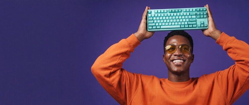 Black man holding computer keyboard on head, young African American developer in orange sweatshirt and sunglasses smiling on purple background with copy space, playful programmer PC tech worker banner