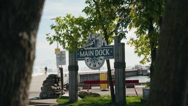 Zoom in on a wooden Main Dock sign with a painted boat, framed by trees, with people, a pier, and Gimli harbor in the background