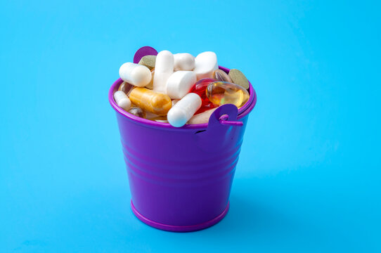 Small purple metal bucket overflowing with mixed pills, capsules, tablets and vitamin supplements on a blue background, concept for medication, pharmacy, healthcare and dietary nutrition
