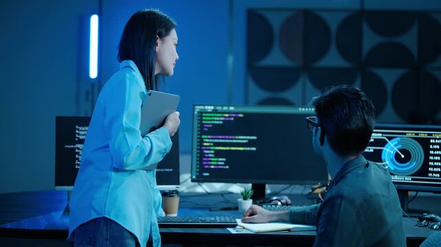 Two Asian software engineers collaborate in a dark modern office. Male programmer points at code on the computer screen, explaining data to his female IT colleague holding a tablet. Teamwork concept.