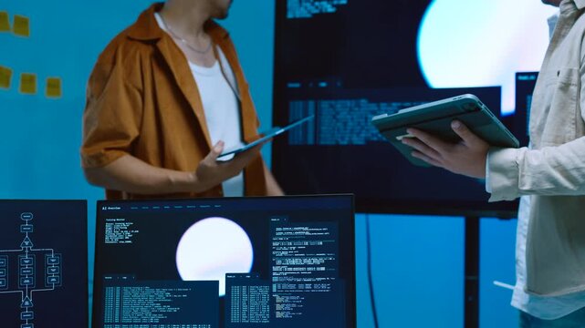 Two young male developers looking at large monitor screen with AI code backend data and talking about system optimization while holding digital tablets in office
