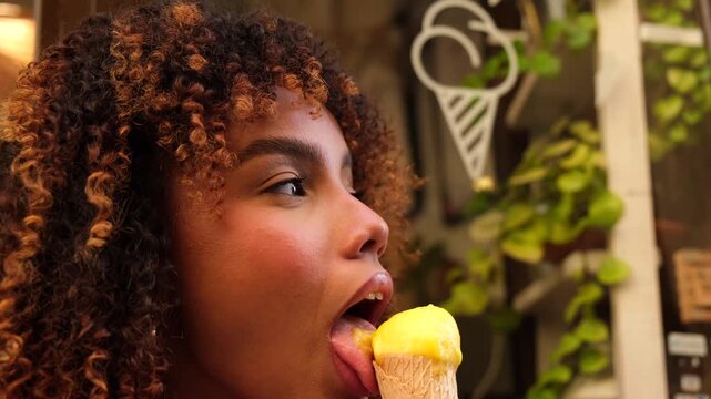Happy african american woman with gorgeous curly hair enjoying a yellow sorbet in a waffle cone, sensually licking the sweet dessert while standing outdoors near a gelateria