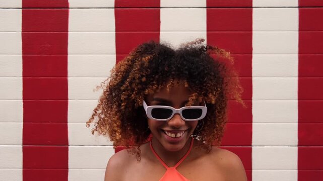 Beautiful african american woman with curly hair dancing and making funny faces in front of a striped wall, she is wearing trendy white sunglasses and enjoying the summer day with joy