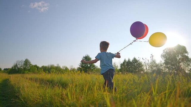 Boy runs through meadow holding colorful balloons. Child plays with balloon bundle at sunset. Kid running in field. Boy carries balloons while playing. Child runs outdoor with colorful balloon.