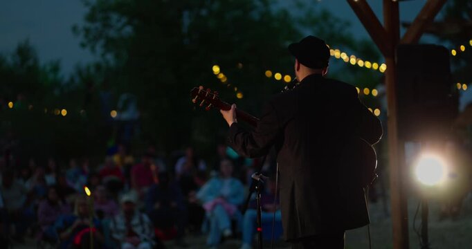 Male guitarist playing acoustic on outdoor evening stage with string lights