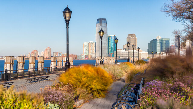 Urban waterfront park walkway with benches and lampposts overlooking the Jersey City skyline, illustrating travel and leisure in Lower Manhattan, New York City, USA.
